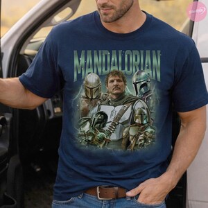 May include: Navy blue t-shirt featuring a graphic of characters from The Mandalorian. The design includes the text "MANDALORIAN" in green, with images of the Mandalorian, Grogu, and other characters. The shirt has a crew neck and short sleeves.
