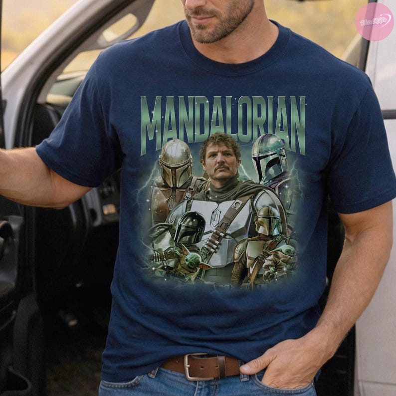 May include: Navy blue t-shirt featuring a graphic of characters from The Mandalorian. The design includes the text "MANDALORIAN" in green, with images of the Mandalorian, Grogu, and other characters. The shirt has a crew neck and short sleeves.