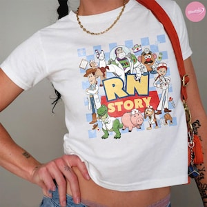 Puede incluir: Una camiseta blanca de manga corta con un estampado gr&aacute;fico de varios personajes de dibujos animados vestidos como profesionales m&eacute;dicos. El texto RN STORY est&aacute; impreso en letras grandes amarillas y rojas. Se lleva una cadena dorada alrededor del cuello.