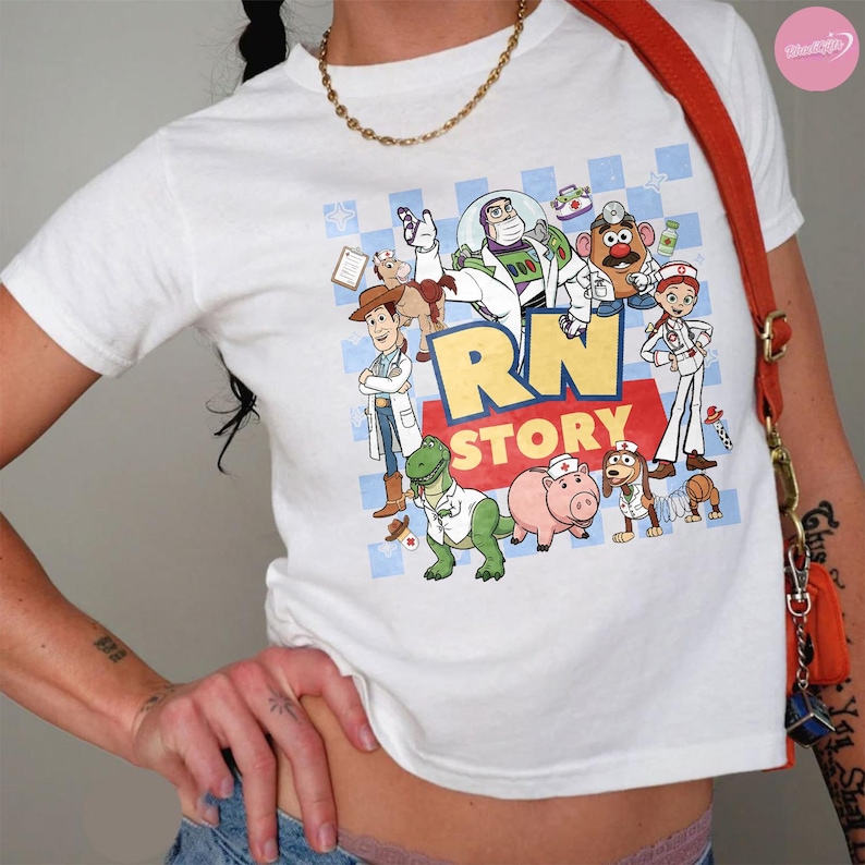 Puede incluir: Una camiseta blanca de manga corta con un estampado gr&aacute;fico de varios personajes de dibujos animados vestidos como profesionales m&eacute;dicos. El texto RN STORY est&aacute; impreso en letras grandes amarillas y rojas. Se lleva una cadena dorada alrededor del cuello.