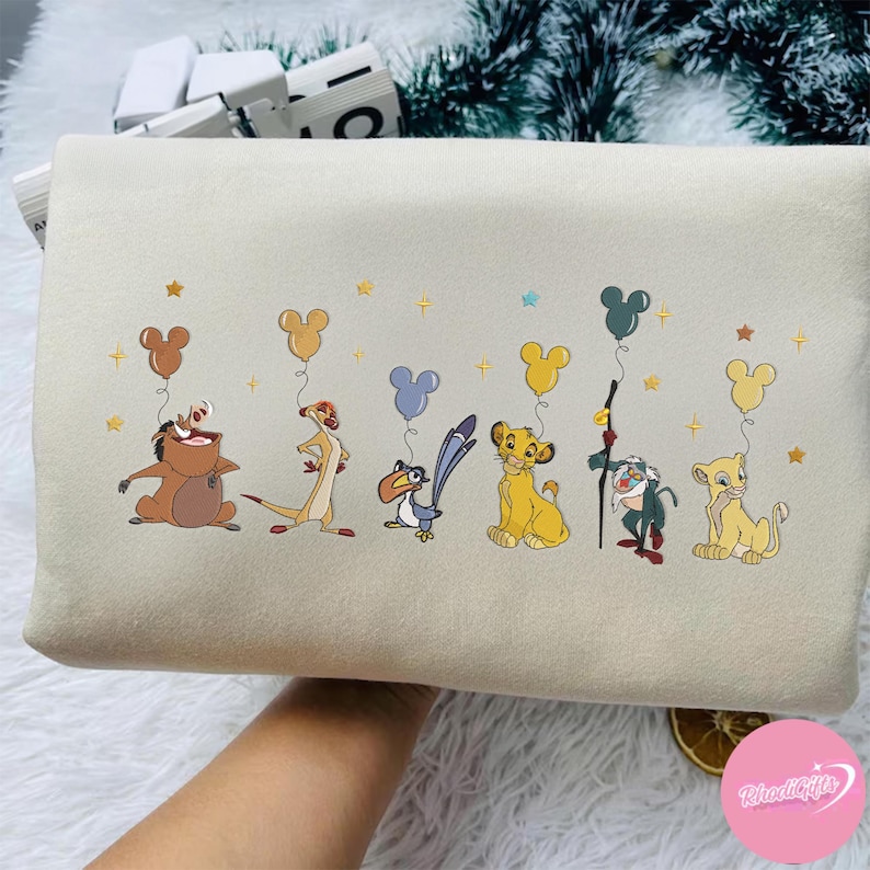 K&ouml;nnte beinhalten: Cremefarbener Kissenbezug mit gestickten Charakteren aus Der K&ouml;nig der L&ouml;wen, darunter Timon, Pumbaa, Simba und Zazu. Jeder Charakter h&auml;lt einen Mickey Mouse-f&ouml;rmigen Ballon. Goldene Sterne und Mickey-Luftballons sind &uuml;ber das Design verstreut.