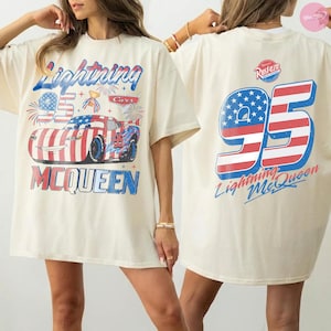 May include: Cream-colored t-shirt featuring a graphic of Lightning McQueen from the Cars movie. The front design includes the number 95, fireworks, and the text "Lightning McQueen". The back of the shirt also displays the number 95 and the text "Lightning McQueen".