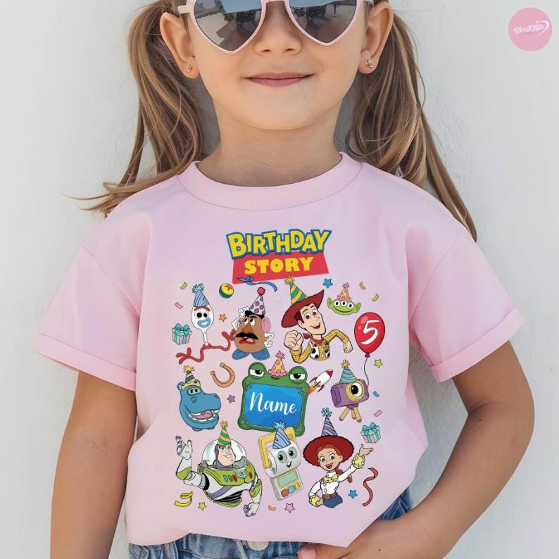 May include: A light pink short-sleeve t-shirt featuring a colorful graphic print of various animated characters wearing party hats. The text "BIRTHDAY STORY" appears at the top, and a central rectangular frame contains the word "Name".