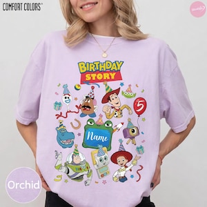 May include: A light purple short-sleeve t-shirt featuring a graphic print with the text "BIRTHDAY STORY" and "Name" surrounded by various cartoon characters wearing party hats, including a cowboy, space ranger, potato figure, and alien.