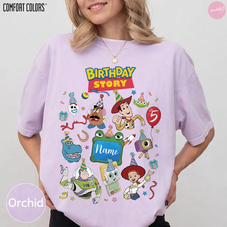 May include: A light purple short-sleeve t-shirt featuring a graphic print with the text "BIRTHDAY STORY" and "Name" surrounded by various cartoon characters wearing party hats, including a cowboy, space ranger, potato figure, and alien.