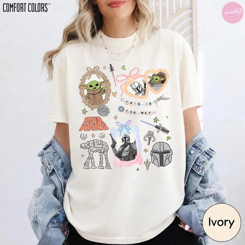 May include: Ivory-colored t-shirt featuring Star Wars-themed graphics. The design includes images of Grogu, the Mandalorian, and other iconic elements. The text "THIS IS THE WAY" is also present. The shirt is a Comfort Colors brand.