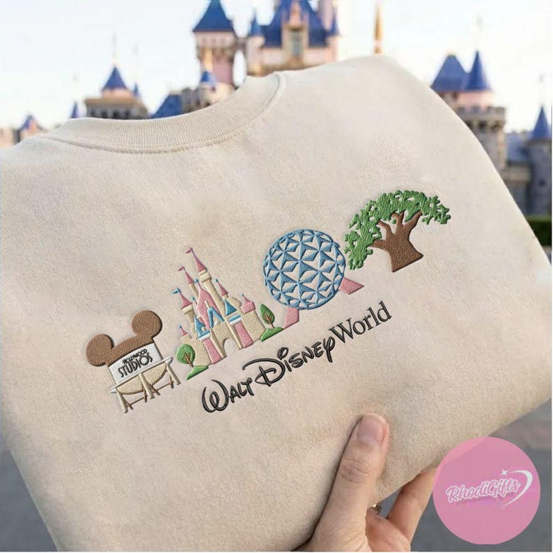 Peut inclure: Sweat-shirt beige avec broderie Walt Disney World. Il pr&eacute;sente une t&ecirc;te de Mickey Mouse marron, un ch&acirc;teau rose et bleu, une sph&egrave;re bleue et blanche et un arbre vert. Les mots "Walt Disney World" sont brod&eacute;s en noir.