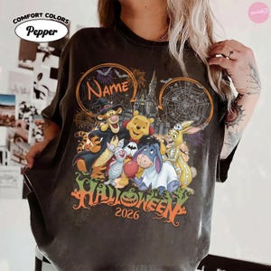 May include: A pepper-colored t-shirt featuring a graphic of Winnie the Pooh characters in Halloween costumes, including Tigger, Piglet, Eeyore, and Rabbit. The design includes a castle, spiderwebs, fireworks, and the text Name, Halloween, and 2026.