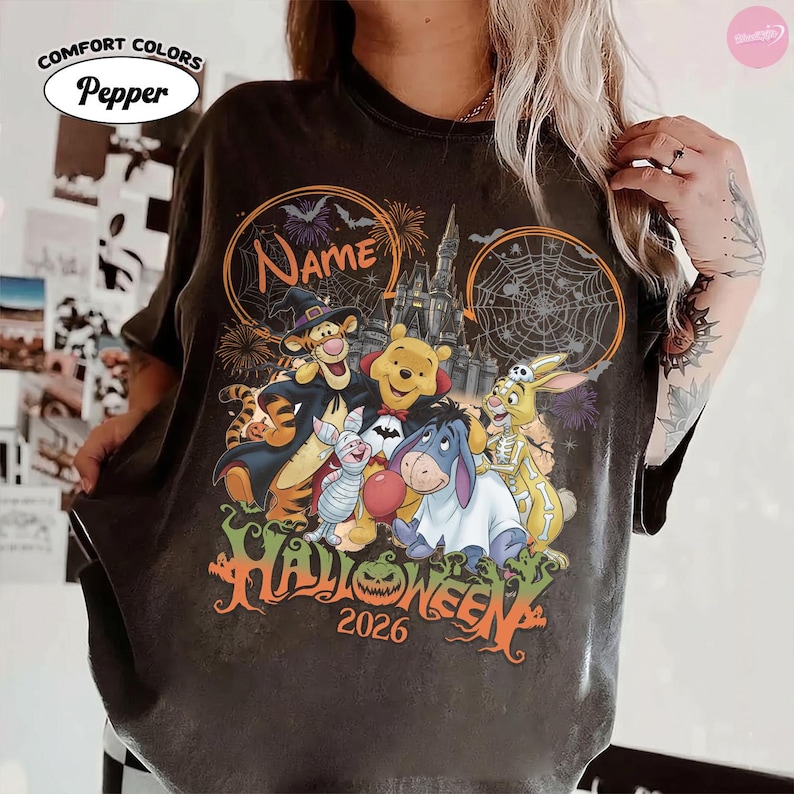 May include: A pepper-colored t-shirt featuring a graphic of Winnie the Pooh characters in Halloween costumes, including Tigger, Piglet, Eeyore, and Rabbit. The design includes a castle, spiderwebs, fireworks, and the text Name, Halloween, and 2026.