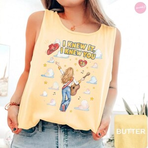May include: A pale yellow sleeveless tank top featuring a graphic illustration of a person with long hair playing an acoustic guitar, surrounded by clouds, stars, and a red cowboy hat. The text reads I KNEW IT, I KNEW YOU with a red heart labeled TS.