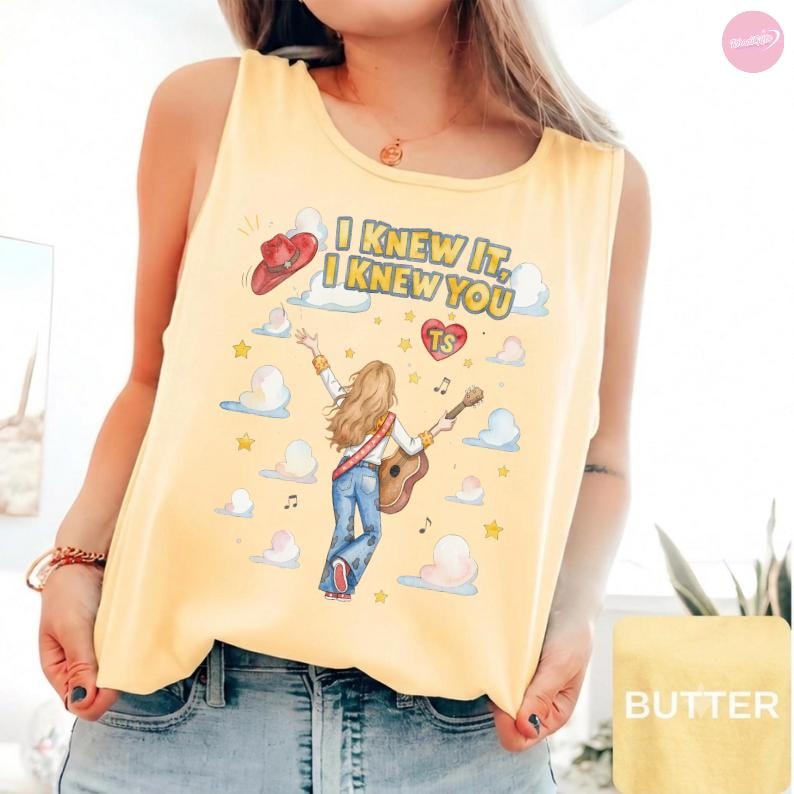 May include: A pale yellow sleeveless tank top featuring a graphic illustration of a person with long hair playing an acoustic guitar, surrounded by clouds, stars, and a red cowboy hat. The text reads I KNEW IT, I KNEW YOU with a red heart labeled TS.