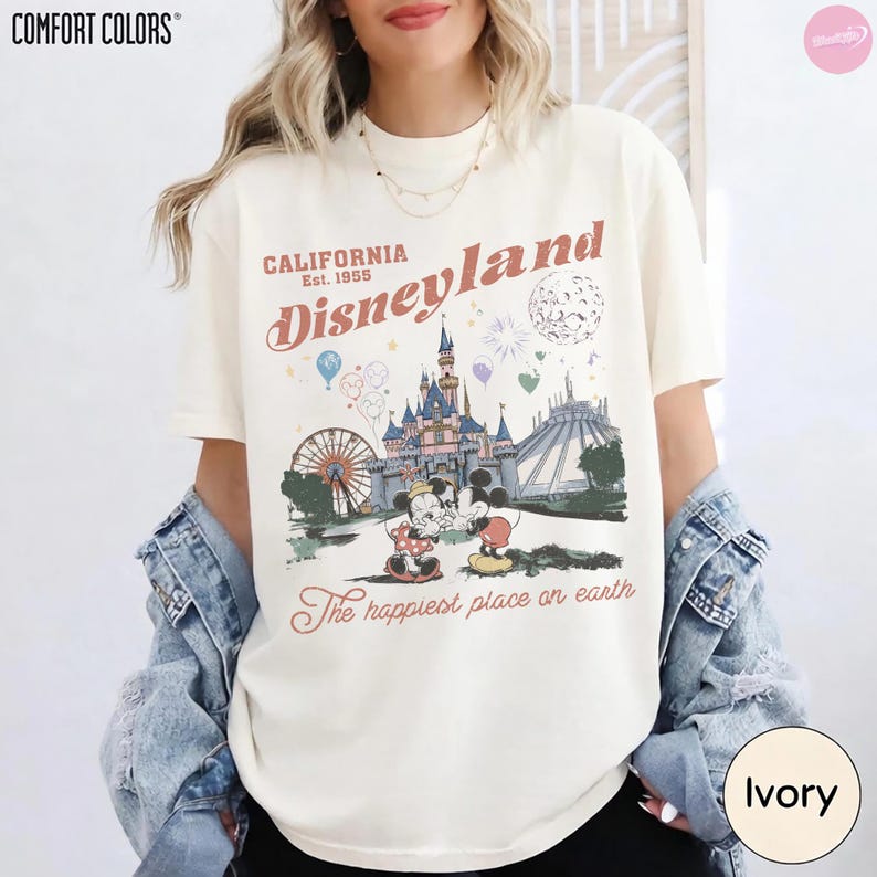 May include: Ivory t-shirt featuring a Disneyland graphic. The design includes a castle, Ferris wheel, and Mickey and Minnie Mouse. Text reads "California Disneyland Est. 1955" and "The happiest place on earth."