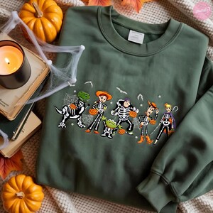 May include: An olive green crewneck sweatshirt featuring a graphic print of six cartoon characters dressed in skeleton costumes for Halloween. The characters are depicted holding orange pumpkins, surrounded by small white bats and spider web designs.
