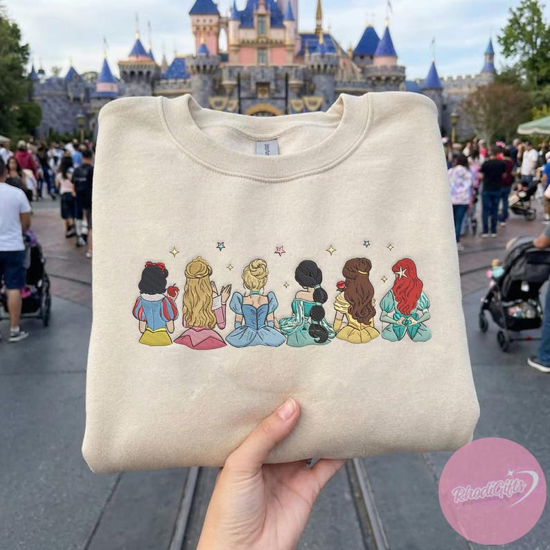 May include: Beige sweatshirt featuring an embroidered design of six cartoon princesses. The princesses are depicted from the back, each with unique hairstyles and dresses in various colors. The sweatshirt is held up in front of a castle.