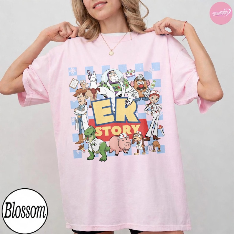 Puede incluir: Una camiseta rosa claro con un estampado gr&aacute;fico de personajes de Toy Story vestidos como personal m&eacute;dico. El dise&ntilde;o incluye a Buzz Lightyear, Woody, Rex, Hamm y el Sr. Patata con el texto ER STORY en letras amarillas grandes sobre una cuadr&iacute;cula azul.