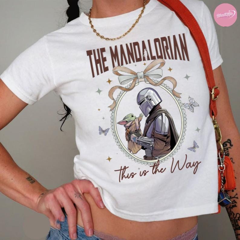 May include: White t-shirt featuring a graphic from The Mandalorian. The design includes the text "THE MANDALORIAN" above an illustration of the Mandalorian and Grogu, surrounded by butterflies and stars. The phrase "This is the Way" is below the illustration.