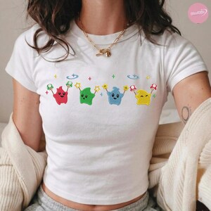 Puede incluir: Camiseta blanca con un dise&ntilde;o bordado de cuatro personajes de dibujos animados en rojo, verde, azul y amarillo. Cada personaje sostiene una estrella y una seta, con nubes y estrellas arriba.