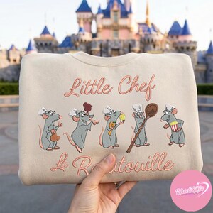 Pode incluir: Camisola bege com ratos de desenho animado bordados usando chap&eacute;us de chef. O texto "Little Chef" e "La Ratatouille" est&aacute; em it&aacute;lico. O fundo mostra um castelo.