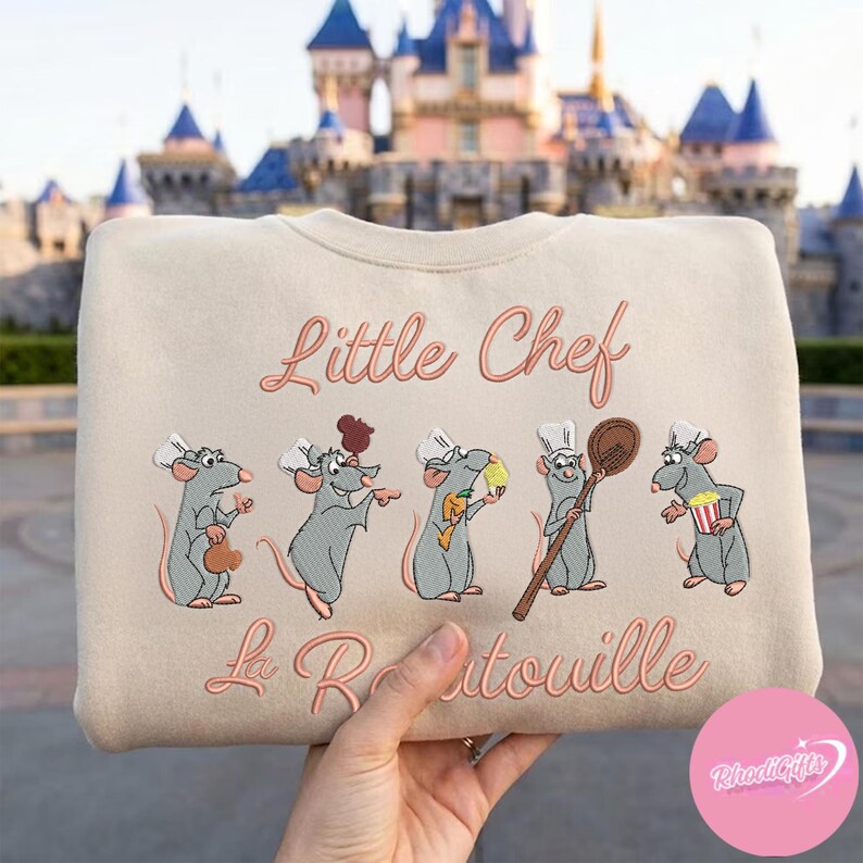 Pode incluir: Camisola bege com ratos de desenho animado bordados usando chap&eacute;us de chef. O texto "Little Chef" e "La Ratatouille" est&aacute; em it&aacute;lico. O fundo mostra um castelo.