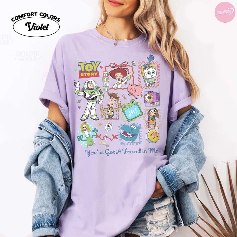 May include: A violet short-sleeve t-shirt featuring a graphic print of Toy Story characters including Buzz Lightyear, Woody, Jessie, and others. The text Toy Story and You've Got A Friend in Me are printed on the front. The shirt is made by Comfort Colors.