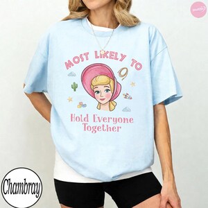 Op de afbeelding: Een lichtblauw t-shirt met korte mouwen en een afbeelding van een blond personage met een roze muts. De tekst "MOST LIKELY TO" staat boven het personage en "Hold Everyone Together" eronder, omringd door kleine iconen van een cactus, sterren en een raket.