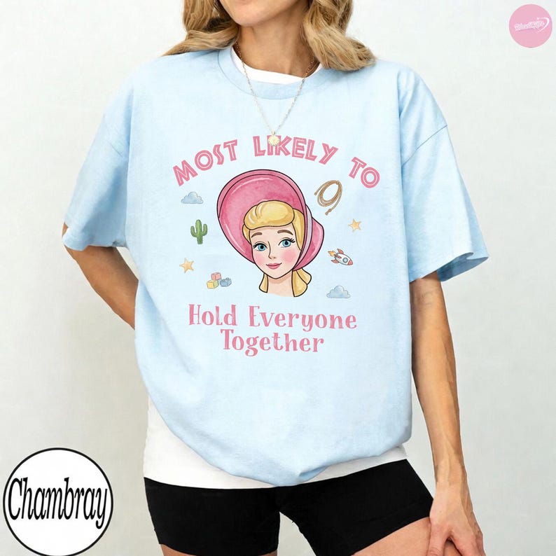 Op de afbeelding: Een lichtblauw t-shirt met korte mouwen en een afbeelding van een blond personage met een roze muts. De tekst "MOST LIKELY TO" staat boven het personage en "Hold Everyone Together" eronder, omringd door kleine iconen van een cactus, sterren en een raket.