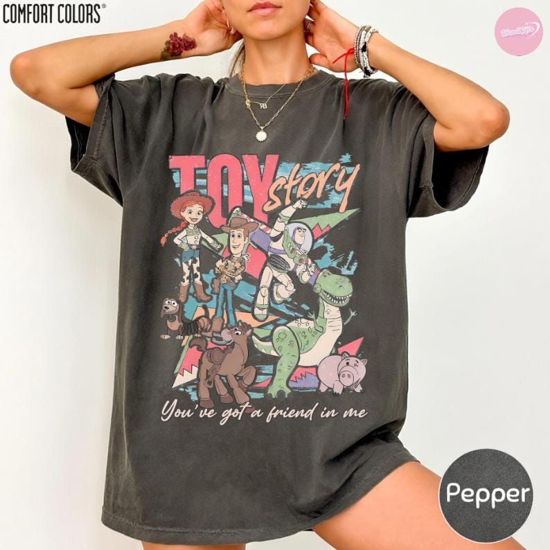Puede incluir: Camiseta color pimienta con un gr&aacute;fico de personajes de la pel&iacute;cula animada "Toy Story". El dise&ntilde;o incluye el texto "Toy Story" en rosa y la frase "You've got a friend in me". La camiseta es de Comfort Colors.
