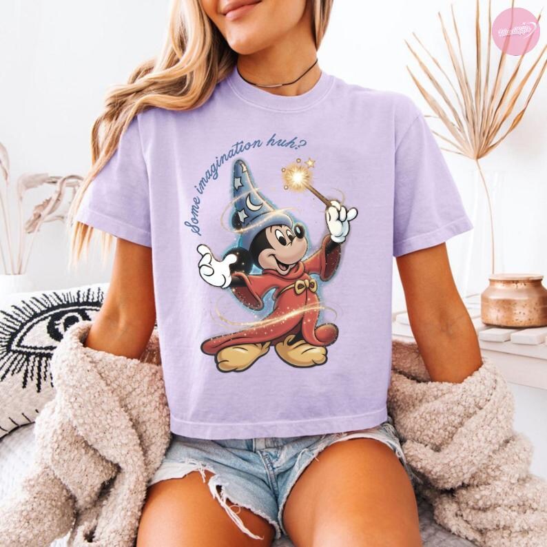 K&ouml;nnte beinhalten: Helllila T-Shirt mit einer Grafik von Mickey Mouse als Zauberer, der einen Zauberstab mit einem funkelnden Stern h&auml;lt. Der Text "some imagination huh?" steht &uuml;ber der Figur. Das Shirt wird mit Jeansshorts getragen.