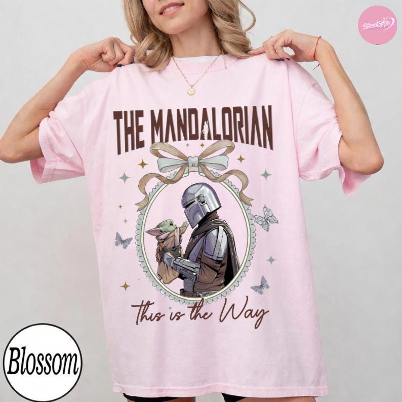 May include: A light pink t-shirt featuring the text "THE MANDALORIAN" above an illustration of the Mandalorian and Grogu. The phrase "This is the Way" is printed below the illustration. The shirt has a round neck and short sleeves.