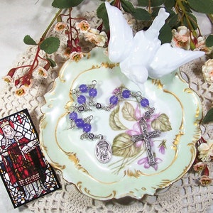 Puede incluir: Un rosario con cuentas moradas y un crucifijo plateado descansa sobre un plato decorativo verde claro con diseños florales. Una figura de paloma blanca y una tarjeta estilo vidriera también están presentes, creando un tema religioso.