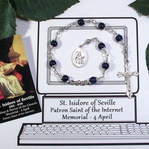 May include: A rosary with dark blue beads and a silver crucifix and pendant. The rosary is displayed on a white background with a drawing of a computer monitor and keyboard. A business card with text about St. Isidore of Seville is also present.