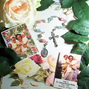 May include: A rosary with pink, white, and blue beads, a silver crucifix, and a pendant with a saint's image. Two cards with a floral background and a picture of a woman holding children are also present. The cards have text.