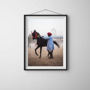 May include: A man in a blue shirt and red turban leads a black horse with a red and white bridle. The horse is walking on a sandy surface. There is a Ferris wheel in the background.