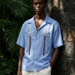 以下が含まれることがあります： 白のヤシの木の刺繍と茶色のビーズのアクセントが付いた、水色の半袖シャツ。 シャツはクラシックな襟とボタンダウンフロントです。 モデルは白いパンツと金のネックレスを着用しています。