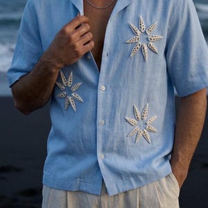 ビーチでの休暇にぴったりの、天然の貝殻があしらわれたメンズライトブルーのリゾートシャツ