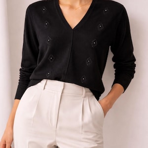 Könnte beinhalten: Schwarzer V-Ausschnitt-Pullover mit rautenförmigen Verzierungen. Dazu eine hellbeige, plissierte Hose. Ein schlichtes, elegantes Outfit für verschiedene Anlässe.