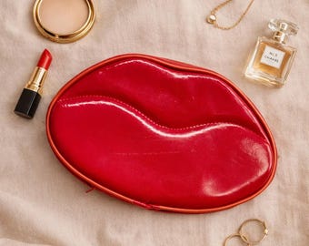 May include: A red lip-shaped cosmetic bag with a zipper, lipstick, compact powder, perfume, a gold necklace, and two gold rings are arranged on a beige surface. The lipstick is red with a black base.