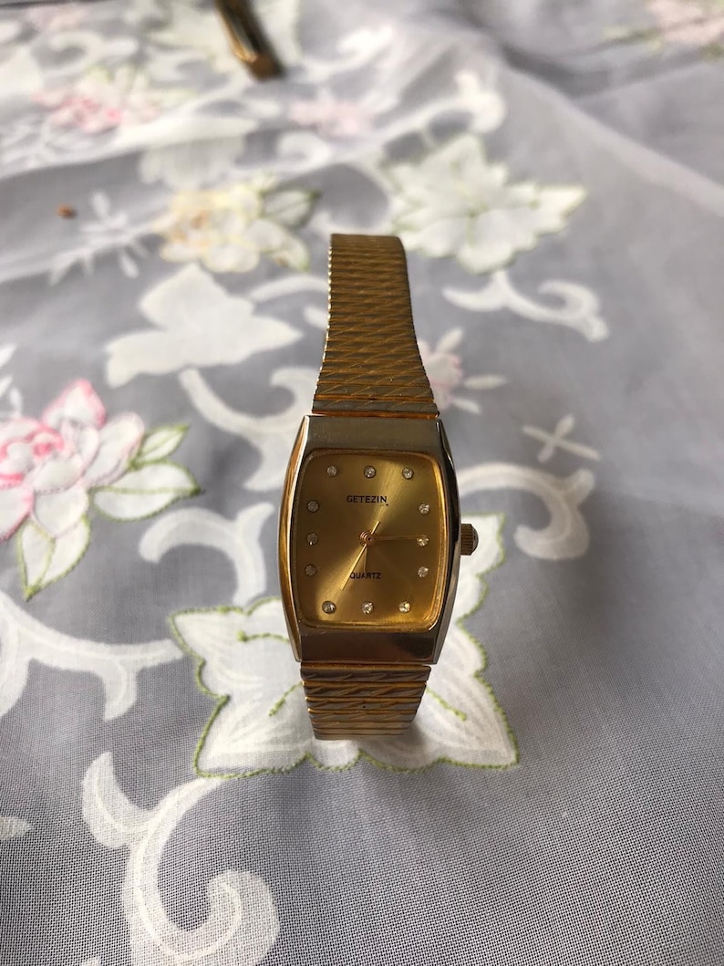 May include: A gold-toned wristwatch with a rectangular face and a gold metal band. The watch face has a gold background with black Roman numeral hour markers and a small seconds dial at the 6 o'clock position. The watch face also has the words "Getezin Quartz" printed on it.