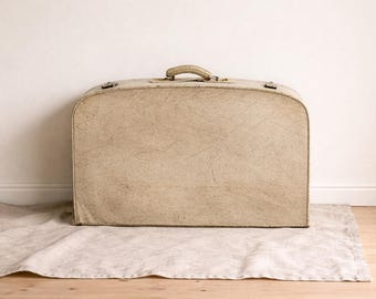 May include: A vintage beige suitcase with a handle and metal clasps rests on a light-colored linen cloth. The suitcase is against a white wall and sits on a wooden floor. The suitcase is a neutral color.
