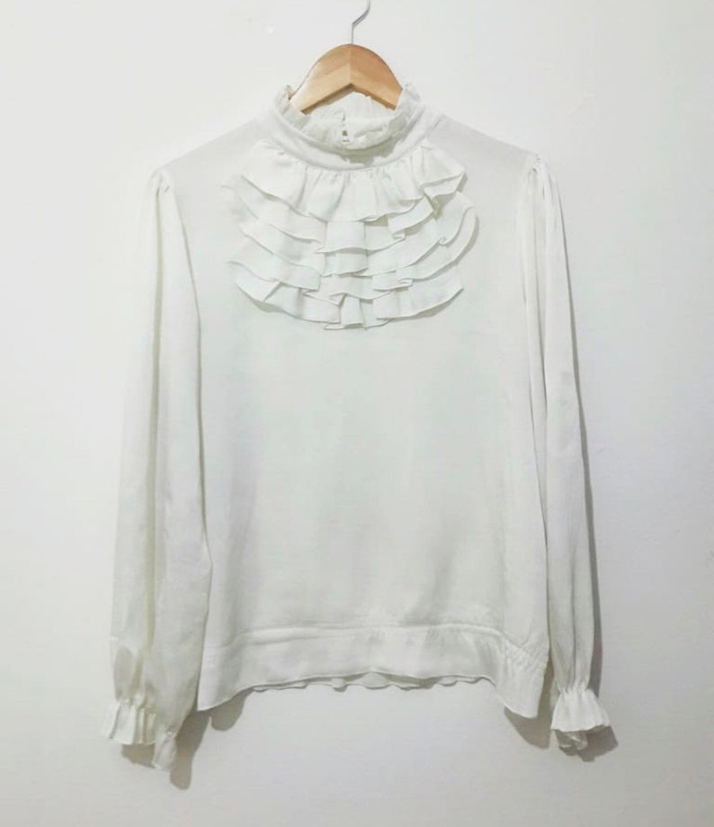 May include: A white long-sleeved blouse with a high ruffled collar. The blouse has a gathered elastic waistband and ruffled cuffs.