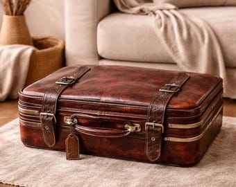 May include: A vintage-style, rectangular brown leather suitcase with two leather straps and metal buckles. The suitcase has a handle and a small brown tag hanging from it. The suitcase is sitting on a neutral-colored rug.