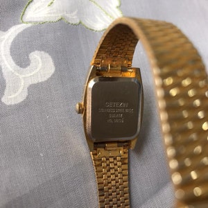 May include: Gold-tone wristwatch with a square face and a gold-tone metal band. The back of the watch has the text "CITIZEN STAINLESS STEEL BACK QUARTZ  WD. 38103".