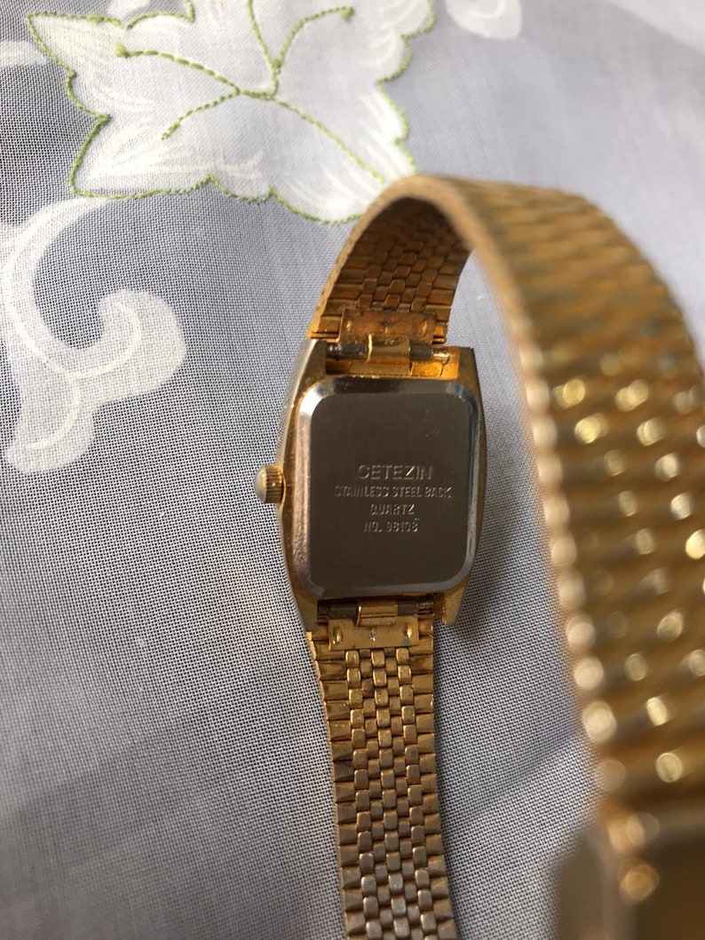 May include: Gold-tone wristwatch with a square face and a gold-tone metal band. The back of the watch has the text "CITIZEN STAINLESS STEEL BACK QUARTZ  WD. 38103".