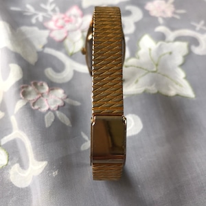 May include: A gold-toned watch band with a textured pattern. The band has a clasp that is visible in the image.