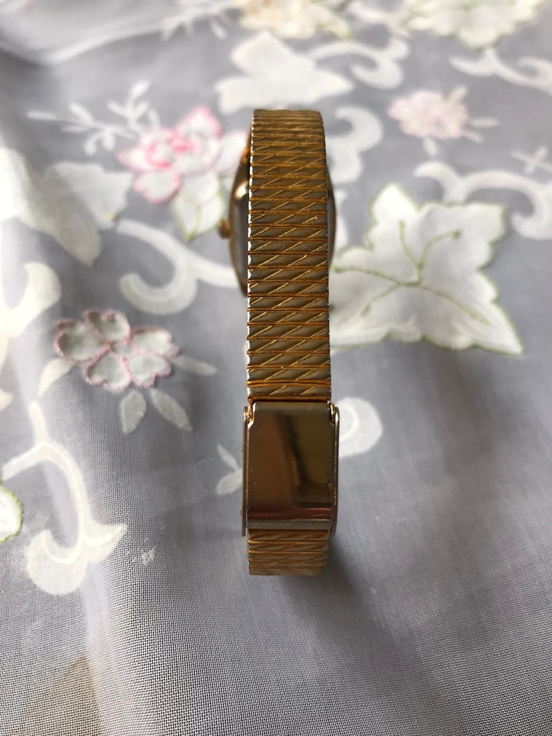 May include: A gold-toned watch band with a textured pattern. The band has a clasp that is visible in the image.