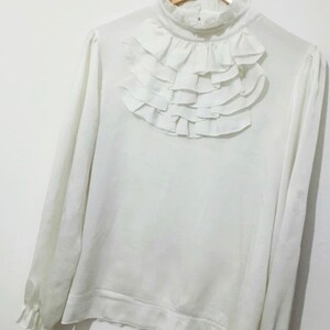 May include: A white long-sleeved blouse with a high ruffled collar. The blouse has a relaxed fit and a slightly gathered hem.