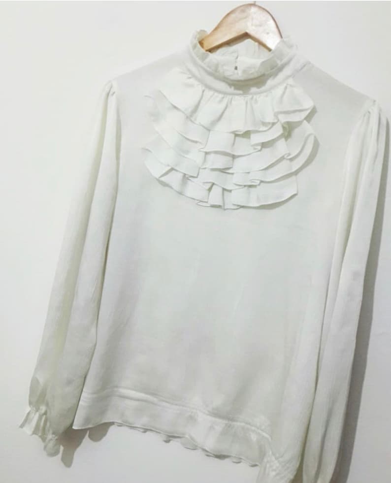 May include: A white long-sleeved blouse with a high ruffled collar. The blouse has a relaxed fit and a slightly gathered hem.