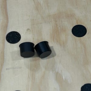May include: Two small, cylindrical black plugs with a smooth, matte finish. Each plug features a flat circular top and a slightly wider rim at the base, designed for insertion into circular openings. The objects are solid black in color.