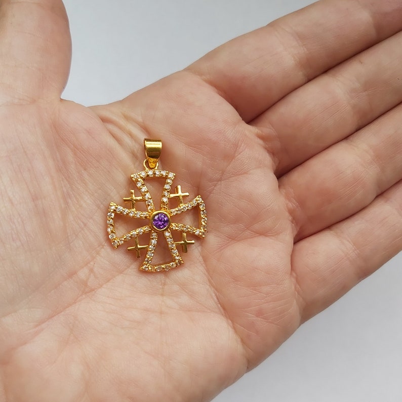 May include: A gold-toned pendant in the shape of a cross with a purple gemstone in the center. The cross is decorated with smaller white gemstones.