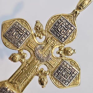 May include: Close-up of a gold-colored cross pendant with intricate detailing. The cross features a central design with a smaller cross and ornate patterns. Silver-colored accents are visible within the design, adding contrast to the gold.